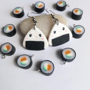 Può includere: Orecchini bianchi e neri a forma di onigiri con ganci argentati, con un volto sorridente e una base di nori di alghe. Intorno agli orecchini ci sono ciondoli a forma di mini rotoli di sushi con ripieni arancioni, verdi e bianchi, su sfondo bianco.