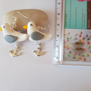 May include: A pair of silver anchor earrings with white and gray seagull charms. The earrings are handmade and feature a delicate design.