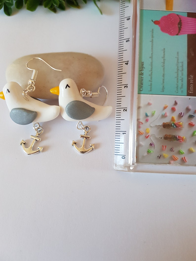 May include: A pair of silver anchor earrings with white and gray seagull charms. The earrings are handmade and feature a delicate design.