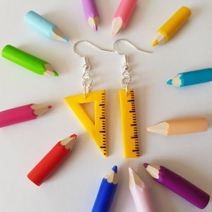 Peut inclure: Une paire de boucles d'oreilles en forme de règle jaune avec des crochets argentés. Les boucles d'oreilles sont entourées d'un cercle de crayons de couleur.
