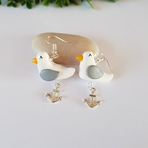 May include: A pair of silver dangle earrings featuring a white seagull with a gray wing and a yellow beak, each with a silver anchor charm hanging below.