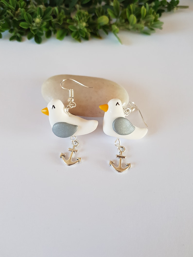 May include: A pair of silver dangle earrings featuring a white seagull with a gray wing and a yellow beak, each with a silver anchor charm hanging below.