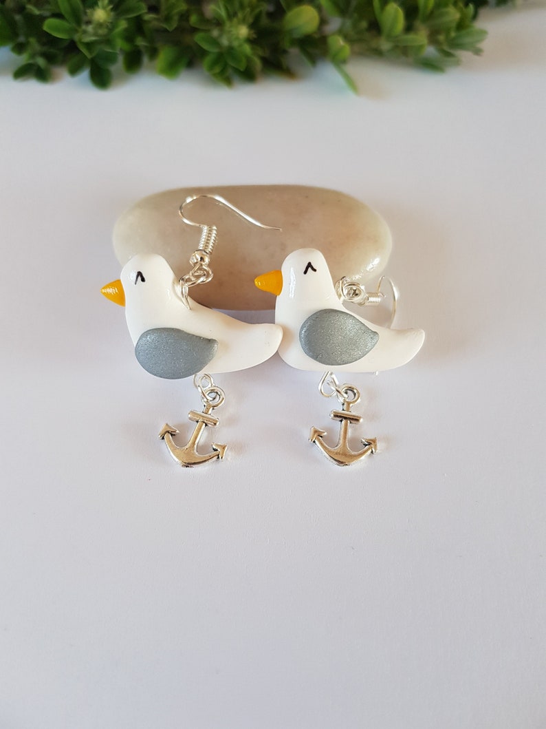 May include: A pair of silver dangle earrings featuring white seagull charms with gray and yellow accents and silver anchor charms.