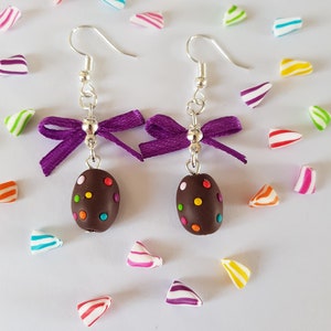 Peut inclure: Une paire de boucles d'oreilles pendantes avec des nœuds en ruban violet et des perles brun chocolat avec des pépites colorées. Les boucles d'oreilles sont sur un fond blanc avec des morceaux de bonbons colorés éparpillés autour.