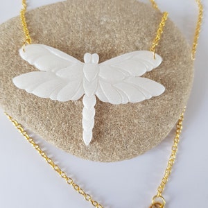 May include: A white, textured dragonfly pendant necklace with a gold chain. The dragonfly is detailed with wings and a body.