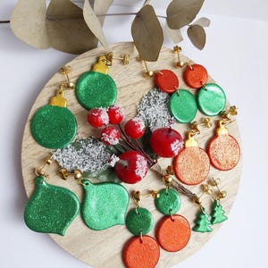 Può includere: Un set di orecchini a tema natalizio con ornamenti verdi e arancioni e un albero di Natale verde. Gli orecchini sono realizzati in pasta polimerica e hanno ganci dorati.