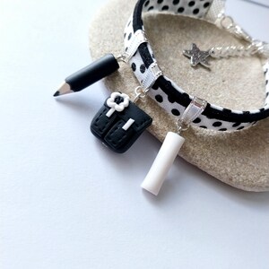 Könnte beinhalten: Ein Armband mit einem schwarz-weiß gepunkteten Band und silberfarbenen Elementen. Das Armband ist mit drei Anhängern versehen: einem schwarzen Stift, einem schwarzen Rucksack mit weißer Blume, einem weißen Zylinder und einem kleinen silbernen Stern.
