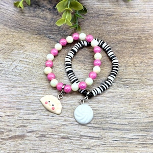 May include: Two beaded bracelets. One bracelet has pink and white beads, the other has black and white striped beads. Each bracelet has a charm. One charm is a white dumpling, the other is a yellow dumpling.