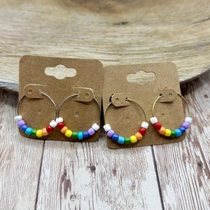May include: Two pairs of hoop earrings with rainbow-colored beads. The earrings are displayed on small brown cardboard earring cards. The hoops are silver-colored and the beads are in a rainbow pattern. The earrings are on a wooden surface.