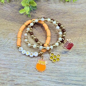 May include: Three beaded bracelets with honey-themed charms. One bracelet features the word "HUNNY" spelled out in white letter beads. Charms include a honey pot, a honeycomb, and a red jar. The bracelets are on a wooden surface.
