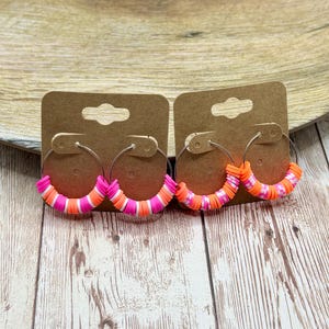 May include: Two pairs of hoop earrings with colorful polymer clay beads. One pair features pink, white, and orange beads, while the other has orange and white beads. The earrings are displayed on brown cardboard earring cards.