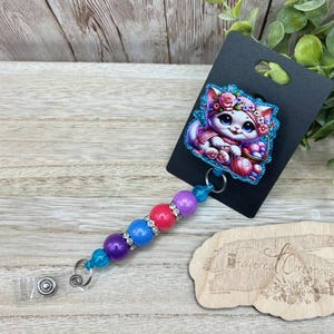 May include: A white cat with pink flowers in its hair and a blue and pink dress is holding a ball of yarn. The cat is on a blue and pink background. The image is on a black card with a white wooden tag that says "Tailored Creations".