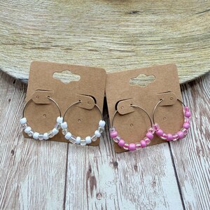 May include: Two pairs of hoop earrings on brown earring cards. One pair features white beads, the other pink. The earrings are displayed on a wooden surface, with a white vase in the background.