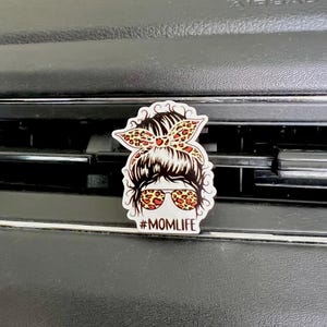 May include: A white car air freshener with a cartoon image of a woman with a messy bun, wearing sunglasses and a leopard print headband. The text "#MOMLIFE" is printed below the image.