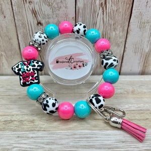 May include: A pink and turquoise beaded bracelet with a cow print charm and a scrub life charm. The bracelet has a silver key chain with a pink tassel.