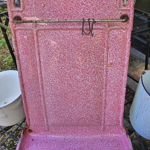 May include: A vintage pink and white speckled enamelware kitchen scale. The scale has a rectangular shape with a curved base and a metal bar for hanging. The enamel has a speckled pattern and shows signs of wear.