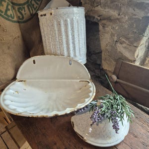 May include: A collection of vintage white enamelware pieces, including a scalloped soap dish, a small wall-mounted dish, and a cylindrical container. The pieces have a distressed, aged appearance with brown edging. A sprig of purple flowers rests on the wooden surface.