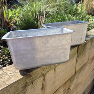 May include: Two rectangular, aluminum baking pans with slightly rounded corners. The pans are sitting on a weathered wooden surface, with green foliage in the background. The pans have a simple, utilitarian design.