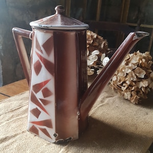 May include: A vintage brown and white enamel coffee pot with a lid and long spout. The pot features a geometric design of brown and red shapes against a white background. The handle and spout are brown. The pot sits on a burlap runner.