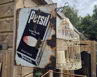 May include: A weathered metal sign for Persil laundry detergent, featuring French text and a black, white, and red design. A decorative birdcage with a candle hangs nearby. The sign is mounted on a wooden structure.