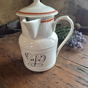 May include: White ceramic coffee pot with a lid and handle. The lid has orange and black stripes. The word "Cafe" is written in black script on the front of the pot. The pot is sitting on a wooden surface.