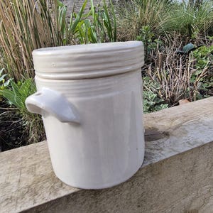May include: A cream-coloured ceramic jar with a lid. The jar has a cylindrical shape with two small handles on the sides. The lid has a ribbed design. The jar is on a wooden surface.