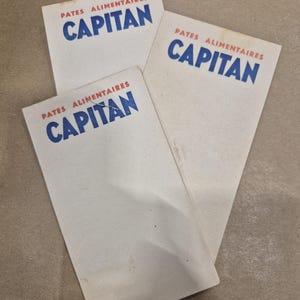 May include: Three white rectangular cards with the text "PATES ALIMENTAIRES CAPITAN" in blue and red. The cards are slightly angled and overlapping, with a neutral background.