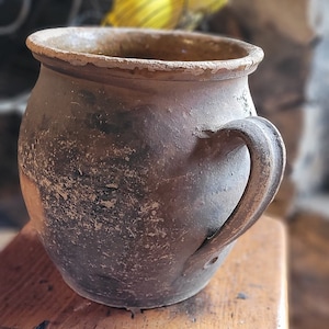May include: A rustic brown clay pot with a single handle. The pot has a worn, textured surface and is slightly out of focus.