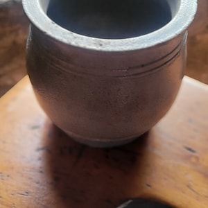 May include: A small, grey ceramic pot with a wide opening and a rounded bottom. The pot is sitting on a wooden surface.