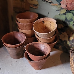 May include: A stack of terracotta clay cups, some are stacked and some are loose, on a white surface. The cups are a light brown color and have a rough texture.
