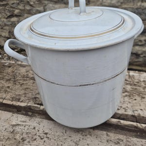 May include: A white enamel chamber pot with a lid and handle. The pot has a rounded shape with a gold band around the middle and on the lid. The handle on the lid has a floral design. The pot is sitting on a wooden surface.