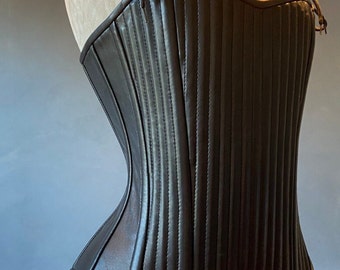 Op de afbeelding: Een zwart leren korset met een veter aan de voorkant en verstelbare bandjes. Het korset heeft een gladde, glanzende afwerking en is ontworpen met verticale panelen.