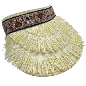 May include: A straw visor with a decorative band. The band features embroidered floral patterns in shades of pink, brown, and gray against a black background. The visor's brim is made of layered, fringed straw.