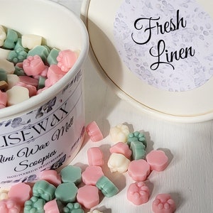 K&ouml;nnte beinhalten: Ein wei&szlig;er Beh&auml;lter gef&uuml;llt mit kleinen, bunten Wachs-Schmelzen in Form von Pfotenabdr&uuml;cken. Der Beh&auml;lter hat ein Etikett mit der Aufschrift "Elise Wax Melts" und "Mini Wax Scoopies". Der Deckel des Beh&auml;lters hat ein Etikett mit der Aufschrift "Frische W&auml;sche".