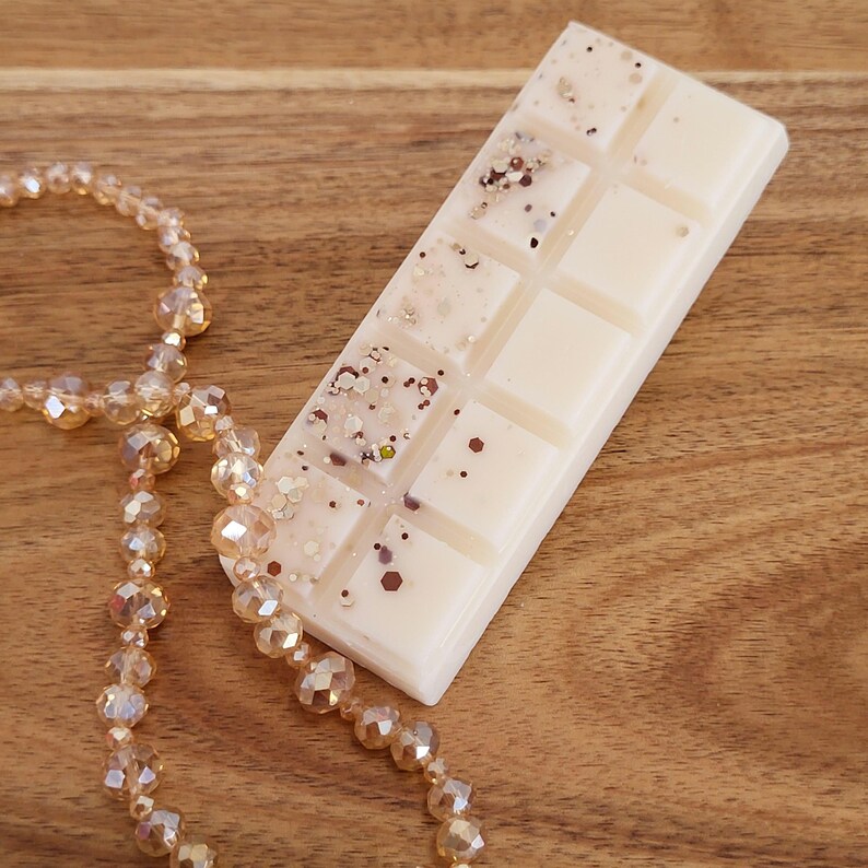 May include: A rectangular, off-white wax melt bar with glitter accents. The bar is divided into ten squares and rests on a wooden surface. A strand of faceted, amber-colored beads is draped nearby, adding a touch of elegance.