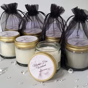 Puede incluir: Seis tarros de vidrio con tapas doradas llenos de velas blancas. Los tarros tienen etiquetas con el texto "Marta's Hen Weekend 30/04/2024" impreso en ellos. Los tarros est&aacute;n dispuestos en una fila sobre una superficie blanca con bolsas de tul negro detr&aacute;s de ellos.
