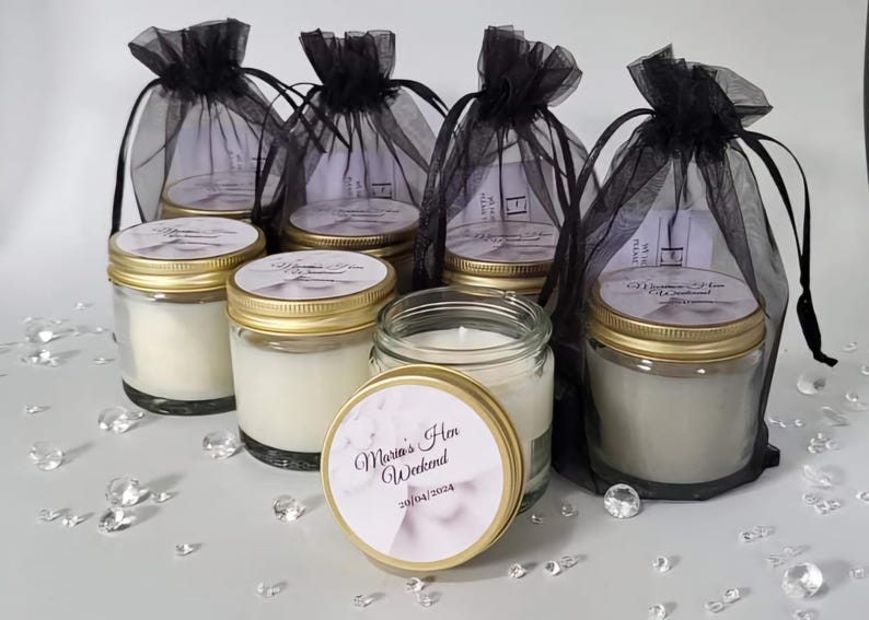 Puede incluir: Seis tarros de vidrio con tapas doradas llenos de velas blancas. Los tarros tienen etiquetas con el texto "Marta's Hen Weekend 30/04/2024" impreso en ellos. Los tarros est&aacute;n dispuestos en una fila sobre una superficie blanca con bolsas de tul negro detr&aacute;s de ellos.