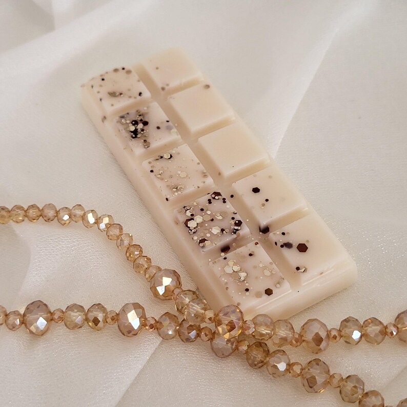 May include: A rectangular wax melt bar with a light beige color, speckled with dark brown and gold glitter. A strand of amber-colored beads is draped in the foreground. The wax melt bar is divided into eight individual squares.