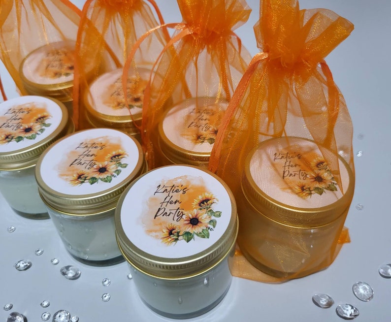 Puede incluir: Conjunto de seis peque&ntilde;os frascos de vidrio con tapas doradas y etiquetas personalizadas. Las etiquetas presentan un dise&ntilde;o de girasol y el texto "Katie's Hen Party". Los frascos est&aacute;n empaquetados en bolsas individuales de tul naranja.