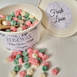 K&ouml;nnte beinhalten: Ein wei&szlig;er Beh&auml;lter gef&uuml;llt mit kleinen, bunten Wachs-Schmelzen in Form von Pfotenabdr&uuml;cken. Der Beh&auml;lter hat ein Blumenmuster und den Text "Elisewax Mini Wax Melt Scoopies" aufgedruckt. Der Deckel des Beh&auml;lters ist wei&szlig; mit einem Blumenmuster und dem Text "Fresh Linen" aufgedruckt. Ein paar Wachsschmelzen sind auf einer wei&szlig;en Oberfl&auml;che verstreut.