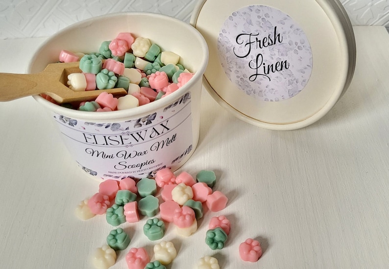 K&ouml;nnte beinhalten: Ein wei&szlig;er Beh&auml;lter gef&uuml;llt mit kleinen, bunten Wachs-Schmelzen in Form von Pfotenabdr&uuml;cken. Der Beh&auml;lter hat ein Blumenmuster und den Text "Elisewax Mini Wax Melt Scoopies" aufgedruckt. Der Deckel des Beh&auml;lters ist wei&szlig; mit einem Blumenmuster und dem Text "Fresh Linen" aufgedruckt. Ein paar Wachsschmelzen sind auf einer wei&szlig;en Oberfl&auml;che verstreut.