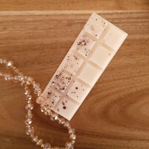 May include: A rectangular, off-white wax melt bar with glitter accents. The bar is divided into ten squares and rests on a wooden surface. A beaded necklace with clear and gold-colored beads is partially visible in the image.
