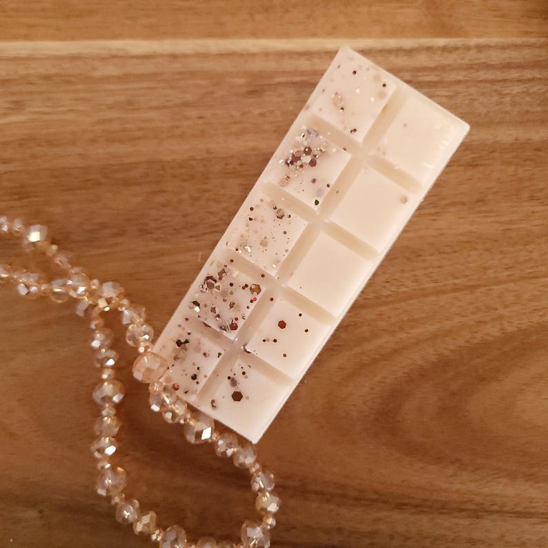May include: A rectangular, off-white wax melt bar with glitter accents. The bar is divided into ten squares and rests on a wooden surface. A beaded necklace with clear and gold-colored beads is partially visible in the image.