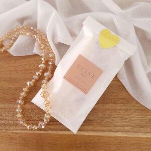 May include: A white paper bag sealed with a gold heart sticker, featuring a pink label that reads "ELISE WAX". A beaded necklace with clear and gold-toned beads is draped on a wooden surface. Soft white fabric is in the background.