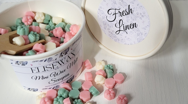 K&ouml;nnte beinhalten: Ein wei&szlig;er Beh&auml;lter gef&uuml;llt mit kleinen, bunten Wachs-Schmelzen in Form von Pfotenabdr&uuml;cken. Der Beh&auml;lter hat ein Etikett mit der Aufschrift "Elise Wax Melts" und "Mini Wax Scoopies". Der Deckel des Beh&auml;lters hat ein Etikett mit der Aufschrift "Frische W&auml;sche".