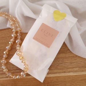May include: A package of Elise Wax, sealed with a yellow heart sticker, rests on a wooden surface. A beaded necklace with amber-colored crystals is draped nearby. The package has a pink label with the text "ELISE WAX" and "Hand Poured | Est. 2021".