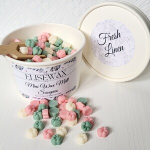 K&ouml;nnte beinhalten: Ein wei&szlig;er Beh&auml;lter gef&uuml;llt mit kleinen, bunten Wachs-Schmelzen in Form von Pfotenabdr&uuml;cken. Der Beh&auml;lter hat ein Etikett mit der Aufschrift "Elisewax Mini Wax Melt Scoopies" und "Hand Poured in Heart of the Home". Der Deckel des Beh&auml;lters hat einen Aufkleber mit der Aufschrift "Fresh Linen".