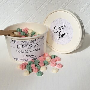 K&ouml;nnte beinhalten: Ein wei&szlig;er Beh&auml;lter gef&uuml;llt mit kleinen, bunten Wachs-Schmelzen in Form von Pfotenabdr&uuml;cken. Der Beh&auml;lter hat ein Etikett mit der Aufschrift "Elisewax Mini Wax Melts Scoopies" und der Deckel tr&auml;gt die Aufschrift "Fresh Linen".