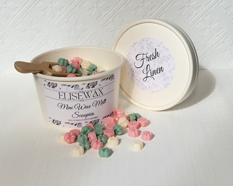 K&ouml;nnte beinhalten: Ein wei&szlig;er Beh&auml;lter gef&uuml;llt mit kleinen, bunten Wachs-Schmelzen in Form von Pfotenabdr&uuml;cken. Der Beh&auml;lter hat ein Etikett mit der Aufschrift "Elisewax Mini Wax Melts Scoopies" und der Deckel tr&auml;gt die Aufschrift "Fresh Linen".