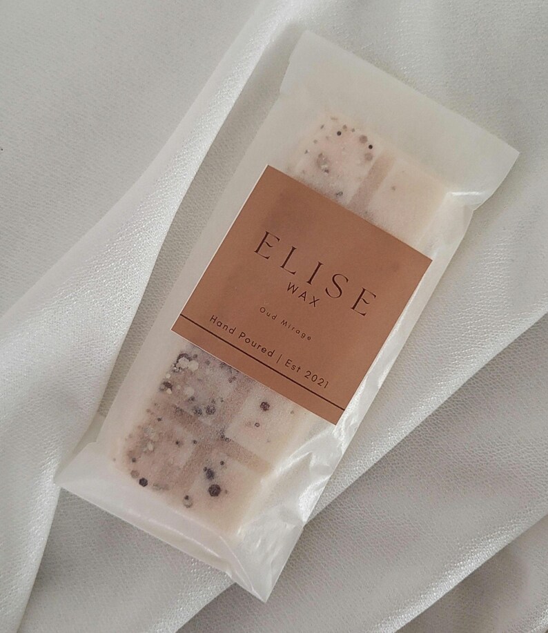 May include: A package of Elise Wax, Oud Mirage scented wax melts. The wax melts are in a clear plastic wrapper with a brown label. The label reads "ELISE WAX" and "Hand Poured | Est 2021". The wax melts have a speckled appearance.